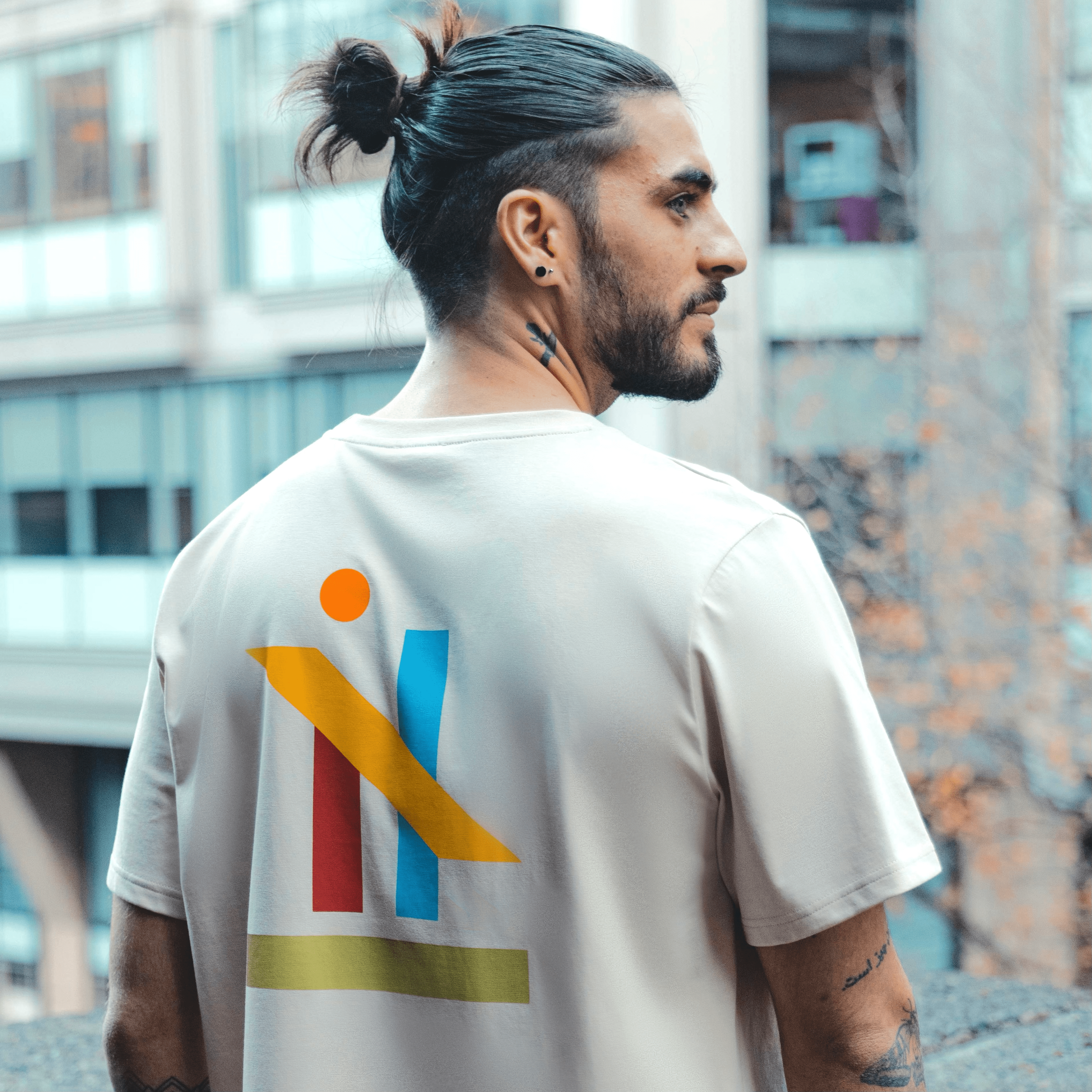 h clothing - male model with tattoos with back to camera wearing sandy tanned tshirt with graphic of colourful geometric shapes