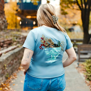h clothing - female model with back to camera wearing blue grey tshirt with graphic of a face and a garden on top of the head