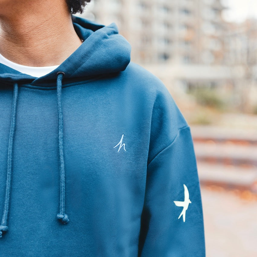 h clothing - close up of male model wearing  sweatshirt hoodie with blue h logo on left breast and coloured bird on left arm