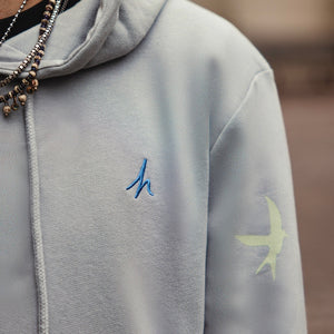 h clothing - close up of male model wearing heather grey hoodie with blue h logo on left breast and green bird on left arm