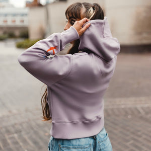 h clothing - female model with back to camera wearing light purple hoodie holding the hood with red bird on left arm