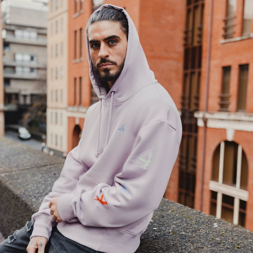 h clothing - male model facing the camera leaning on wall wearing light purple hoodie with colourful birds on left arm