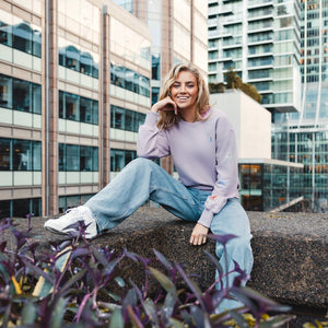 h clothing - female model facing the camera sitting on wall wearing light purple sweatshirt with colourful birds on left arm