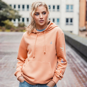 h clothing - female model facing the camera wearing pastel orange hoodie with colourful birds on left arm