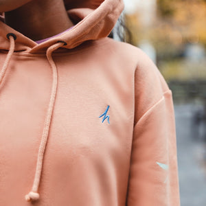 h clothing - close up of female model wearing pastel orange hoodie with blue h logo on left breast