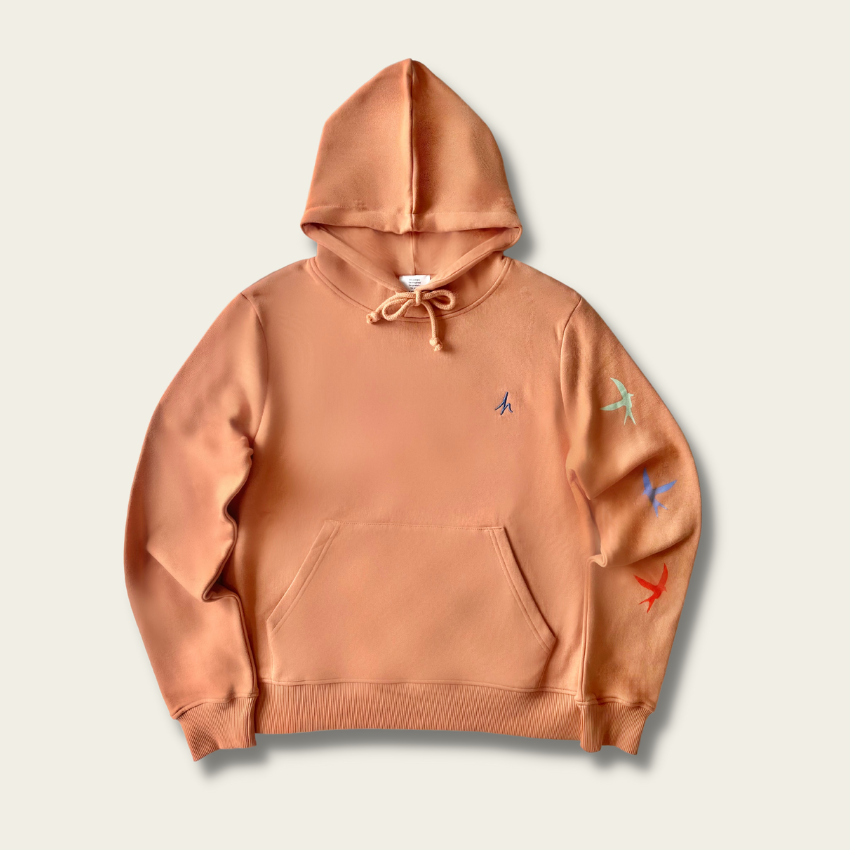 h clothing - flat shot of front of pastel orange hoodie with blue h logo on left breast and colourful birds down the left arm