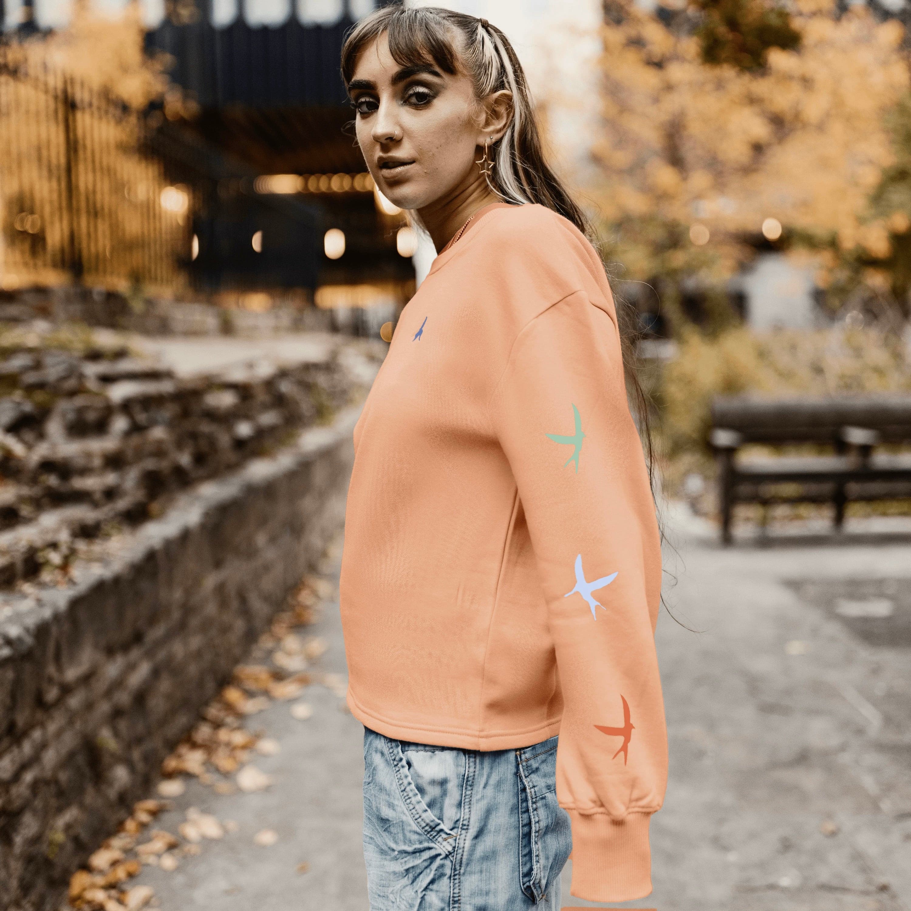 h clothing - female model side on wearing pastel orange sweatshirt with colourful birds going down left arm